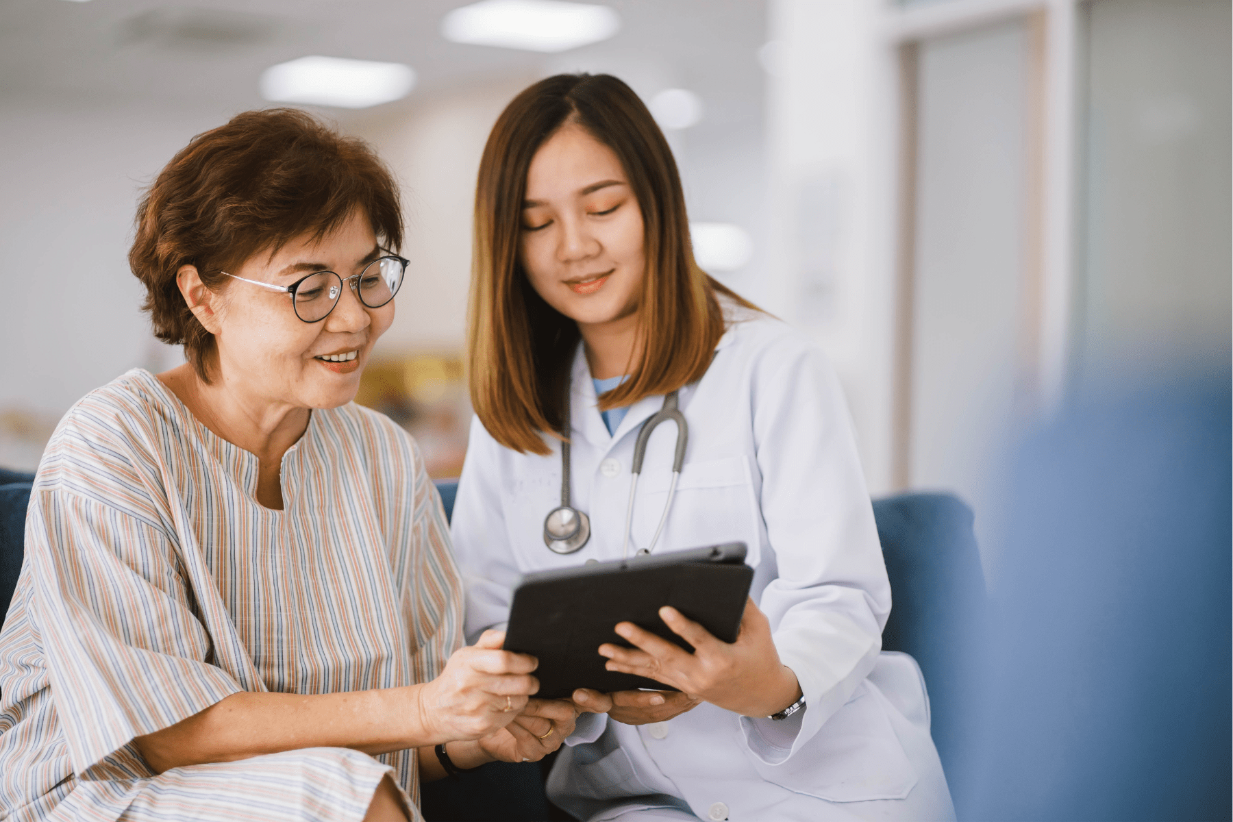 A doctor and patient, both Asian females, sit side by side. The doctor, dressed in a white lab coat with a stethoscope, holds an iPad. Her patient is looking at the iPad with her, and both doctor and patient are smiling.