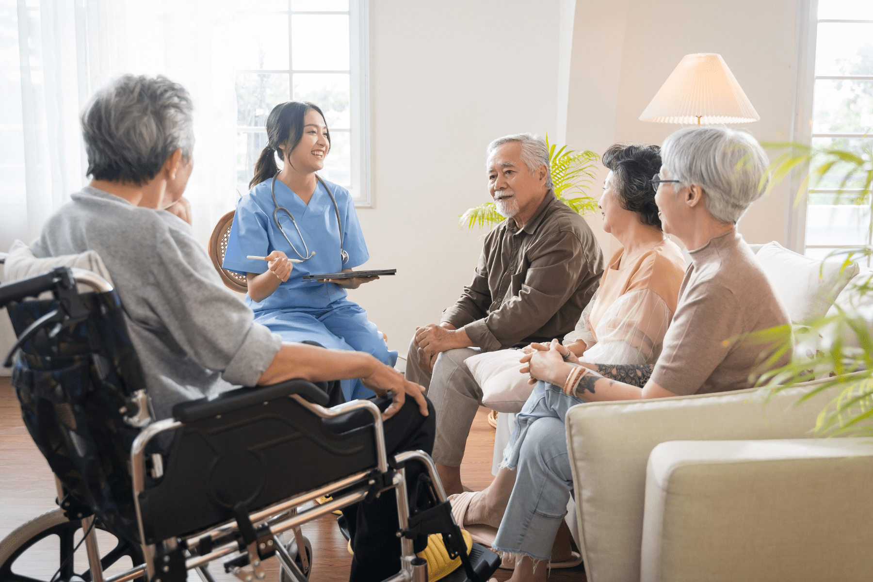 A group of older adults sit in a semi circle. Their attention is directed to a brown-haired nurse in blue scrubs who is addressing the group and similing.