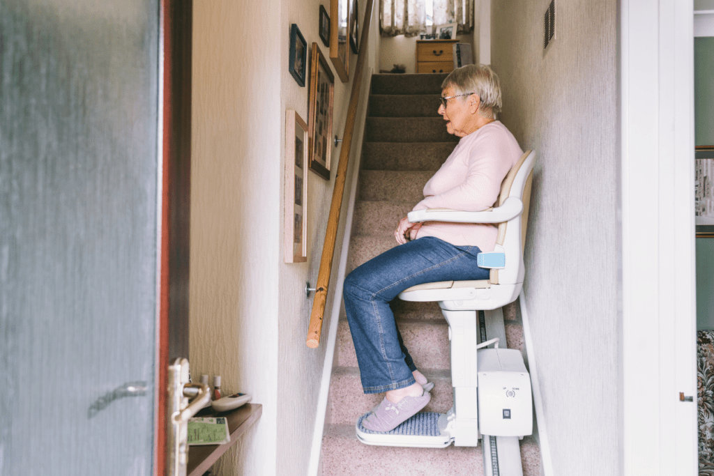 Profile of a Caucasian female in a pink sweather and blue jeans riding a stairlift in her home. She is facing a wall full of photos.