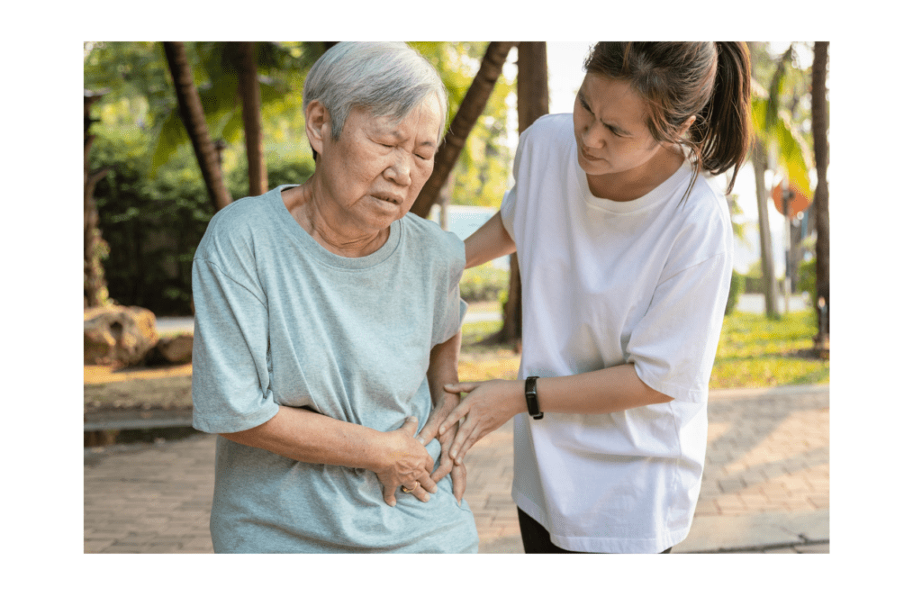 Two Asian women are side by side. The older Asian woman is on the left of the image. She has short grey hair, and a light blue top. She is holding her side in pain. Next to her is a younger Asian woman, with dark hair, a white top, and black slacks. She is assisting the older woman by helping her to walk.