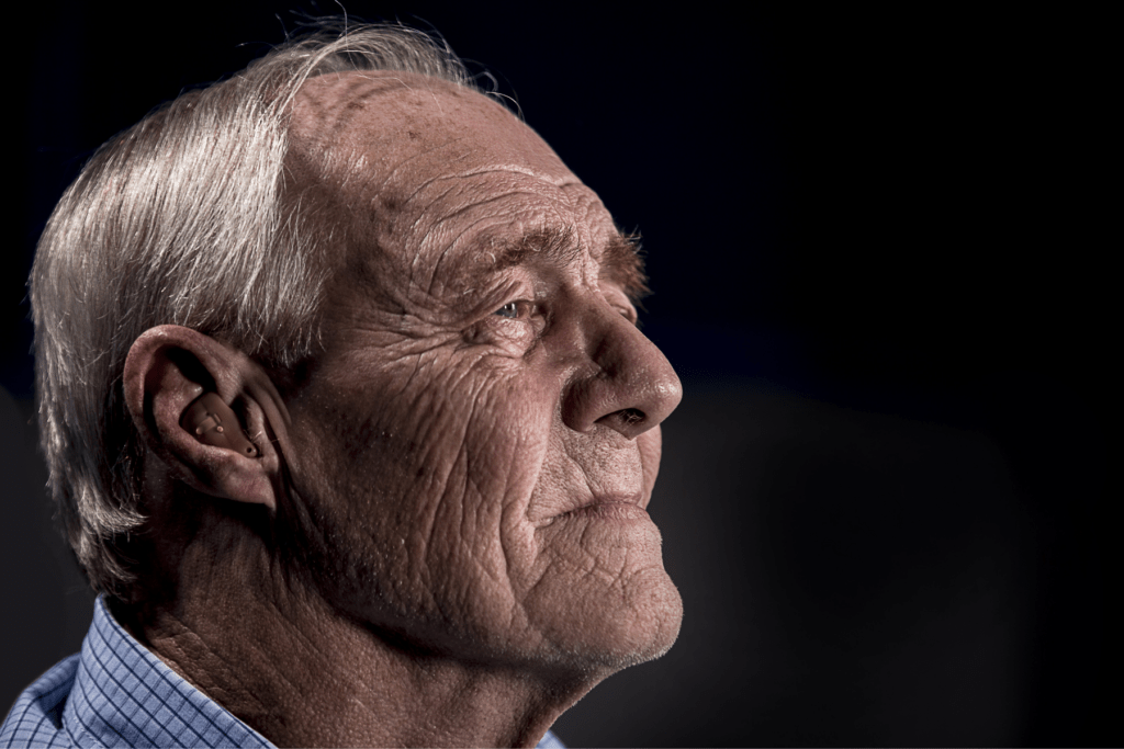 A light-skinned, grey haired man is pictured in profile. He is wearing a hearing aid and a light blue shirt against a dark background. The expression on his face is neutral.