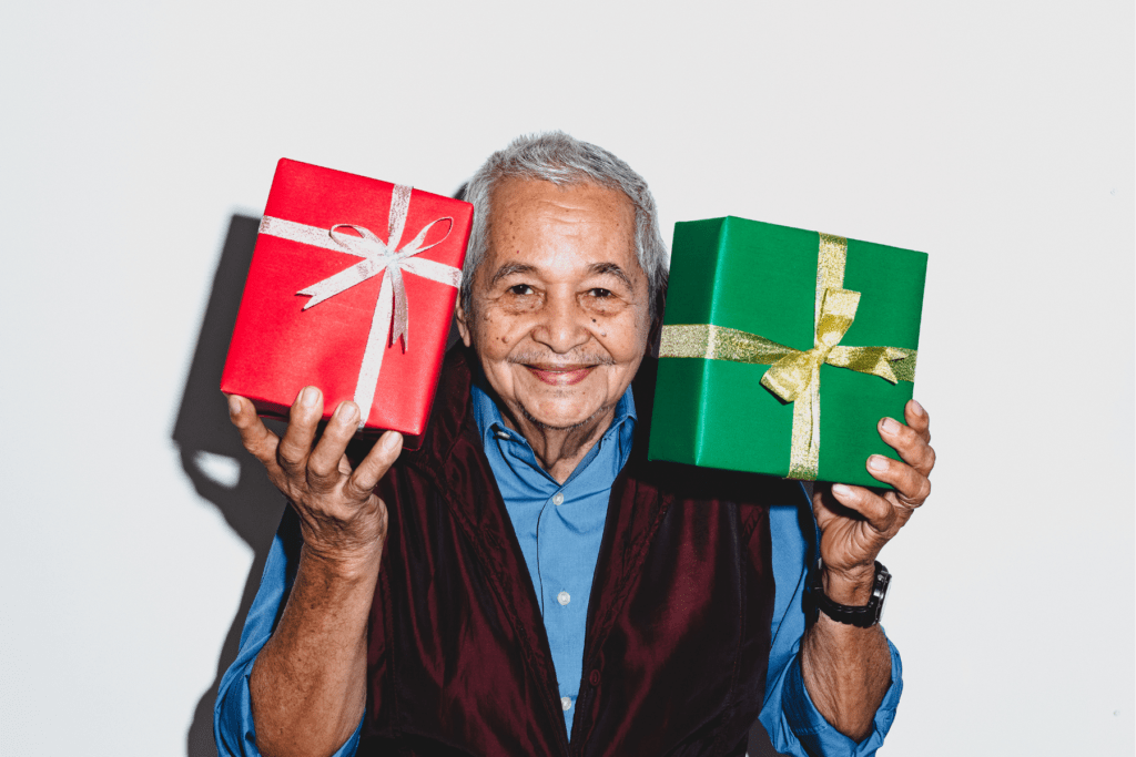 A man of AAPI descent is facing the camera and smiling. He is pictured from the waist up wearing a light blue short-sleeved shirt and a dark blue vest. In one hand, he holds a gift wrapped in one paper, and in his other hand, he holds a gift wrapped in green paper. He is holding each gift up at eye level.