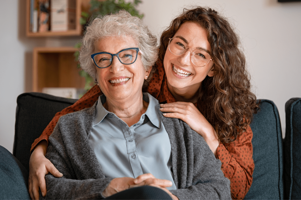 Two women are facing the camera and smiling. The older woman is on the left of the frame and has white hair and blue-framed glasses. She is wearing a grey shirt and dark grey cardigan. Next to her, is a younger woman with brown curly hair, glasses, and is wearing a rust-colored blouse, She is resting her hands on the shoulders of the older woman and is also smiling at the camera.