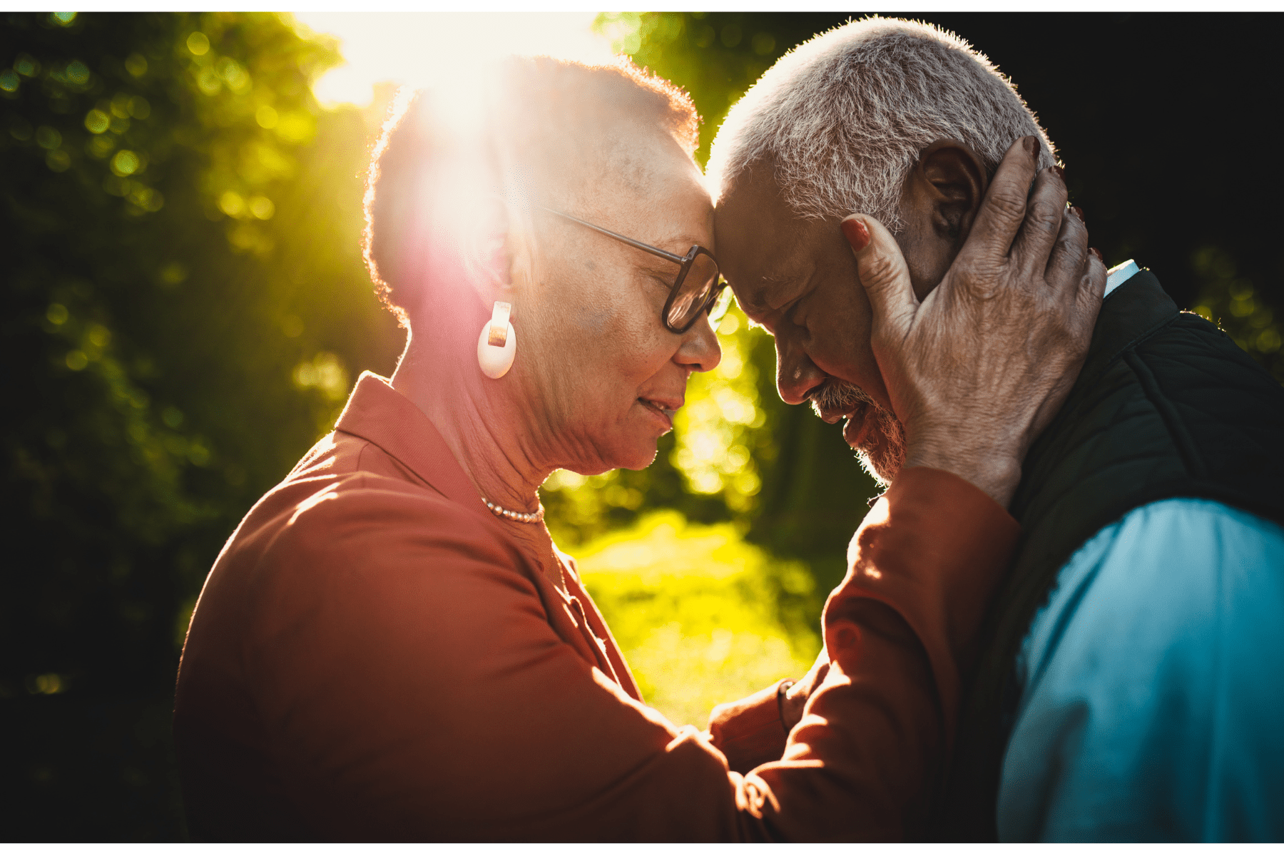 An older Black couple is facing one another, touching foreheads. The woman is in the left side of the image. She is wearing a read top. She has short hair, glasses, and earrings. Her hand is resting gently on the side of the man's face. He is dressed in a blue shirt with a dark blue vest over it. He has short hair. They are both gazing downward. The background is thick green landscaping with the sun peeking through just above the couple.