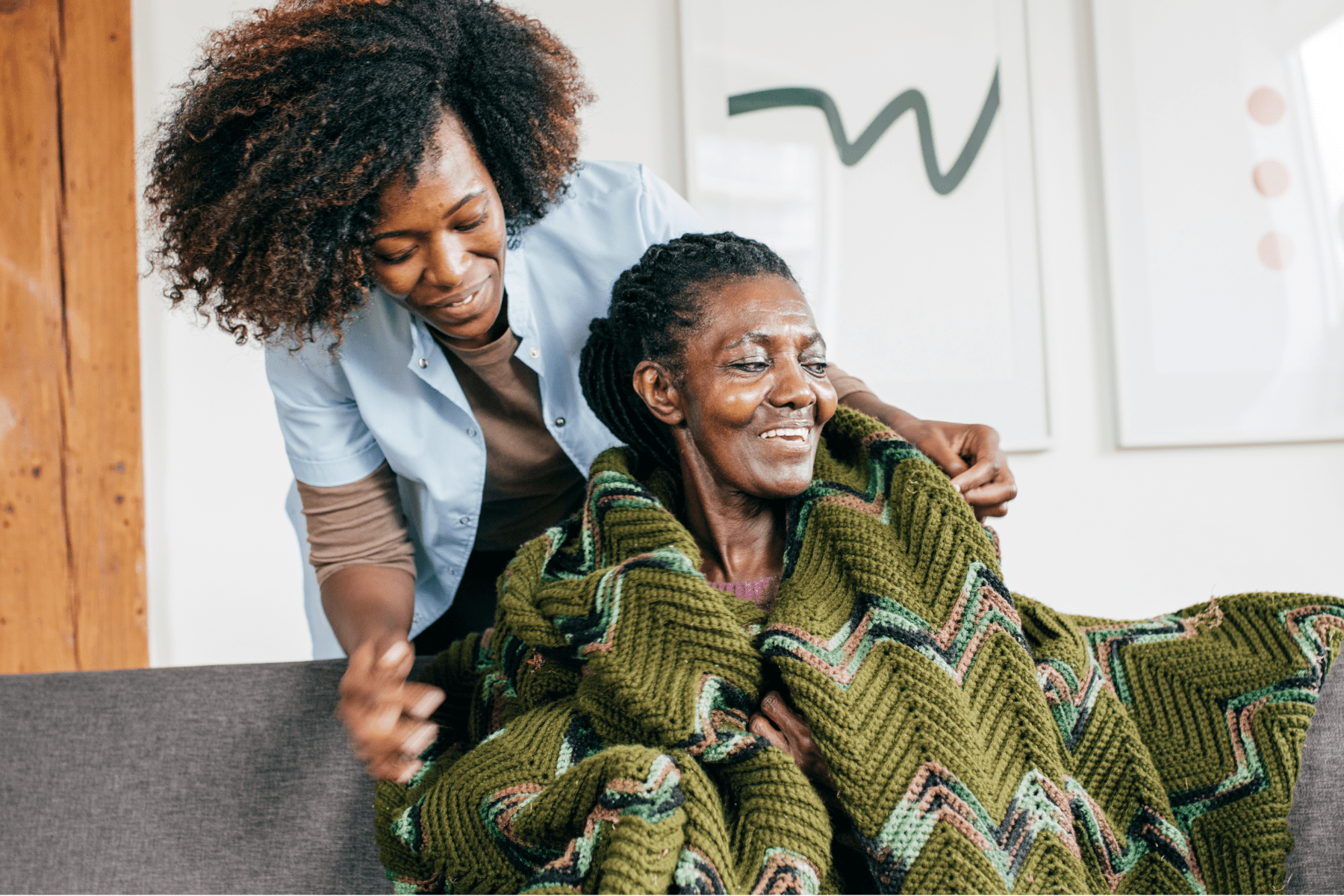 Two Black women are pictured. The younger woman is standing behind the older woman. She has short black hair and is wearing a dark top with a light blue short-sleeved shirt layered over it. She is resting a blanket on the older woman's shoulders. Both women are smiling and are gazing to the side of the camera.