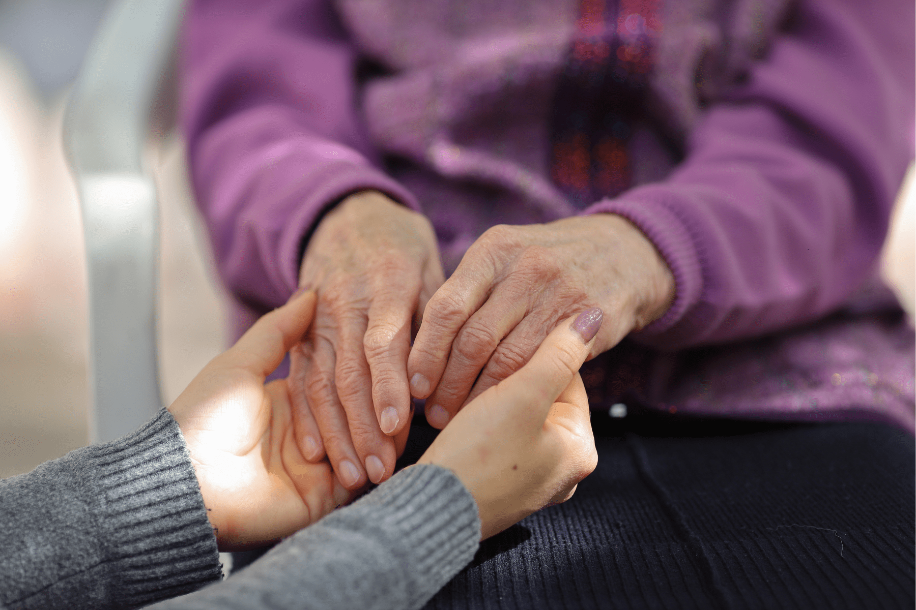 Image shows an older person in a purple sweater whose hands are being held by a young person in a gray shirt and with light pink nail polish. Their faces are not visible.